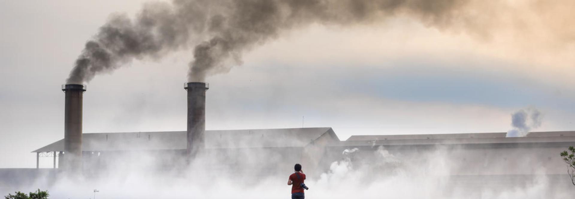 Usine dont 2 cheminées rejettent de la fumée, des déchets au sol et une personne de dos en avant-plan